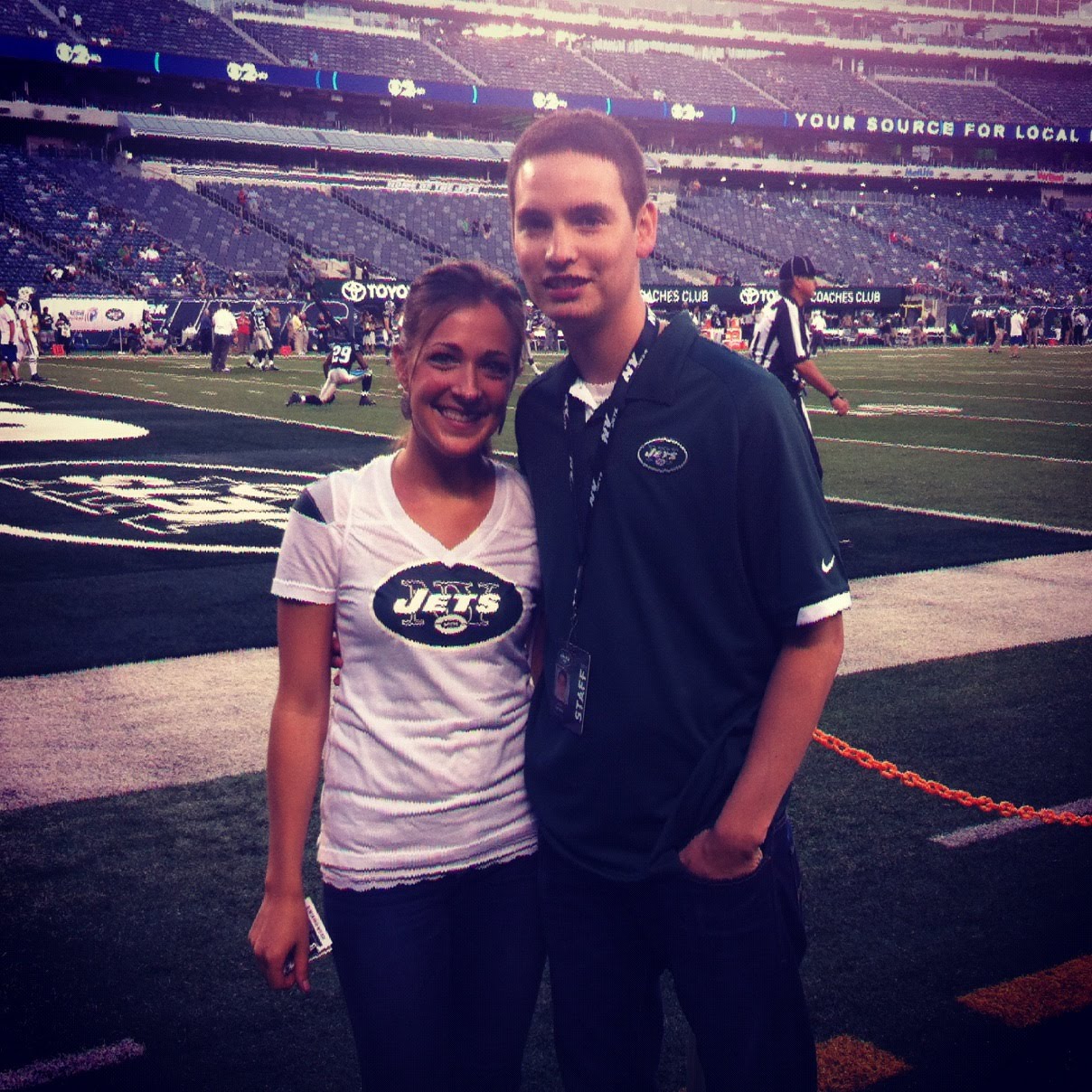 Justin in Jets staff kit with a field credential, next to Steph in a Jets tee, on the field at MetLife before kickoff.