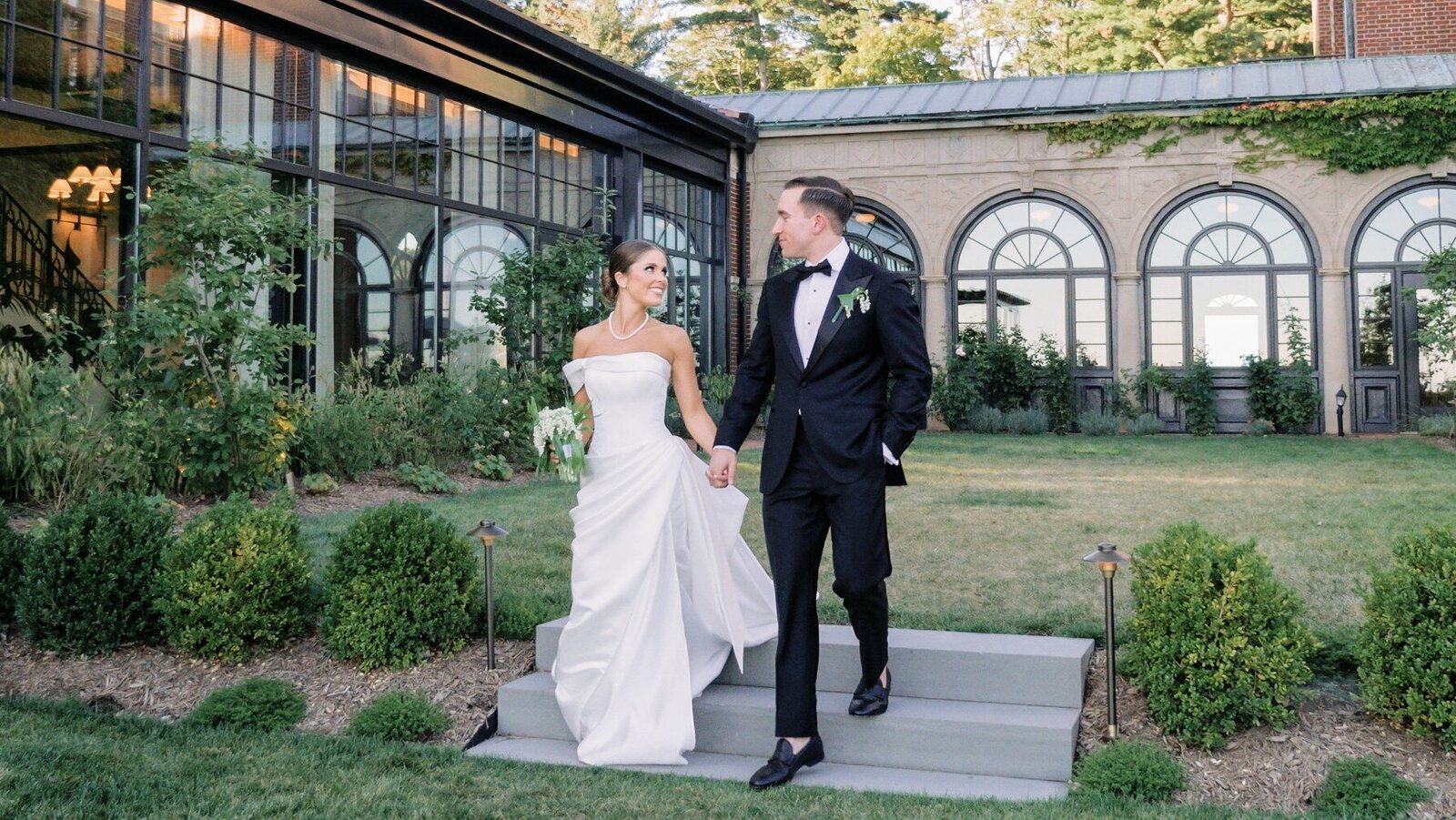A bride and groom hand in hand on the steps outside a glass conservatory at dusk.