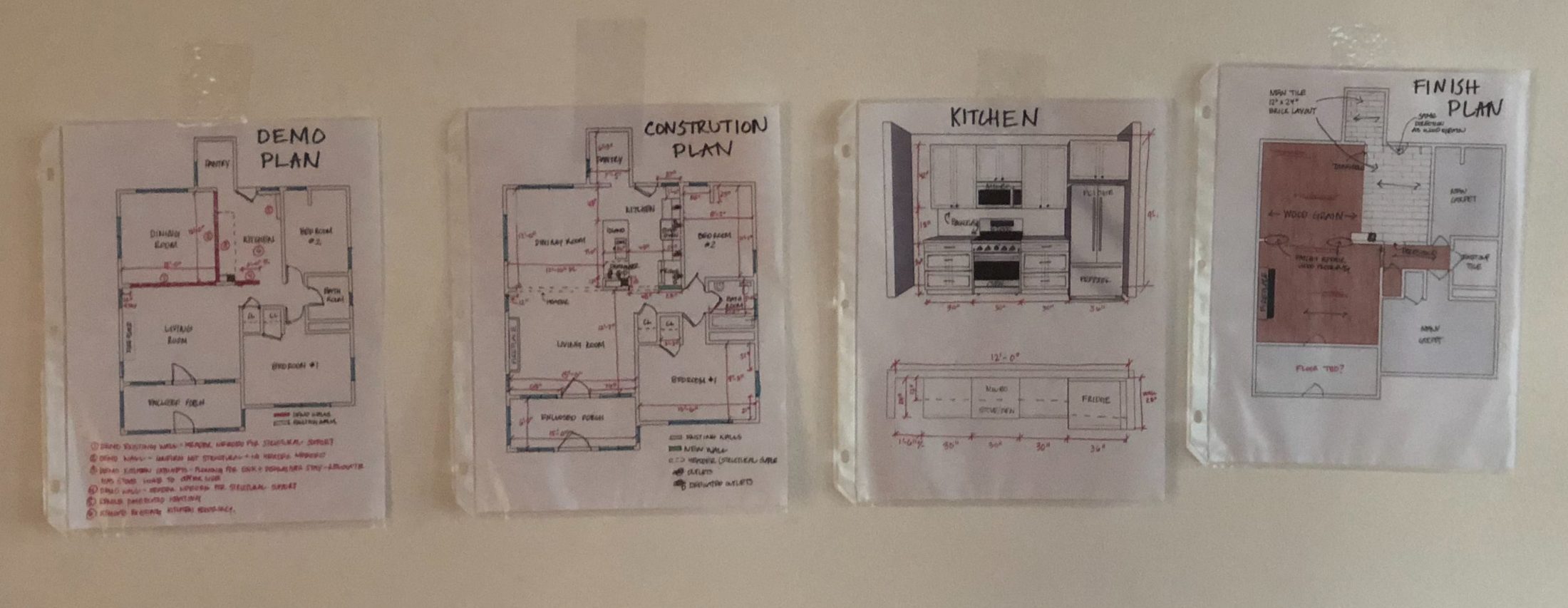 Four of Steph's hand-annotated plans in sheet protectors taped to the wall: demo plan, construction plan, kitchen, finish plan.