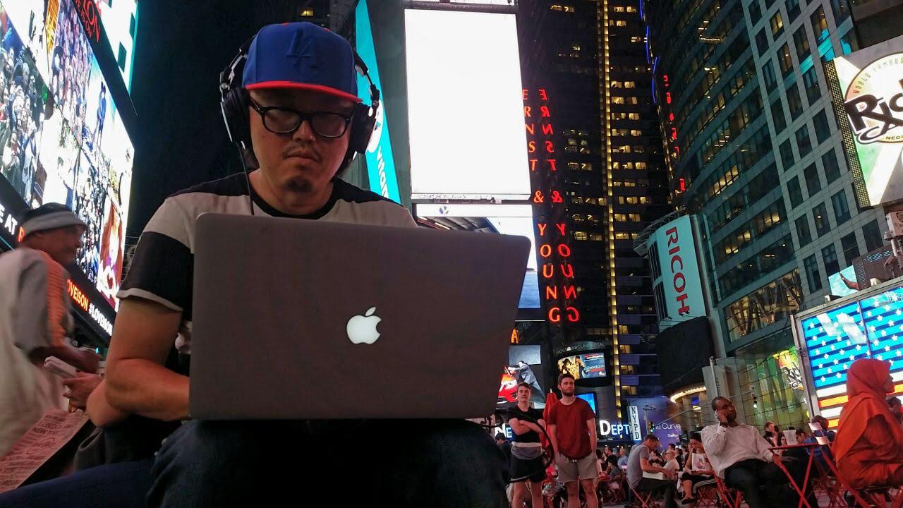 Director Joseph Kahn editing on a laptop in Times Square at night, headphones on, surrounded by neon billboards.