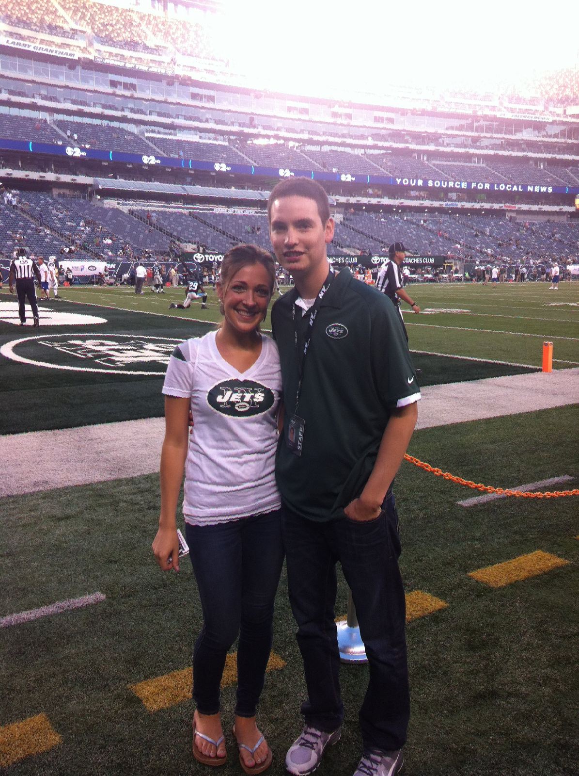 Justin Herrera in a Jets staff polo with a credential lanyard, standing with Steph on the field at MetLife Stadium before a game.