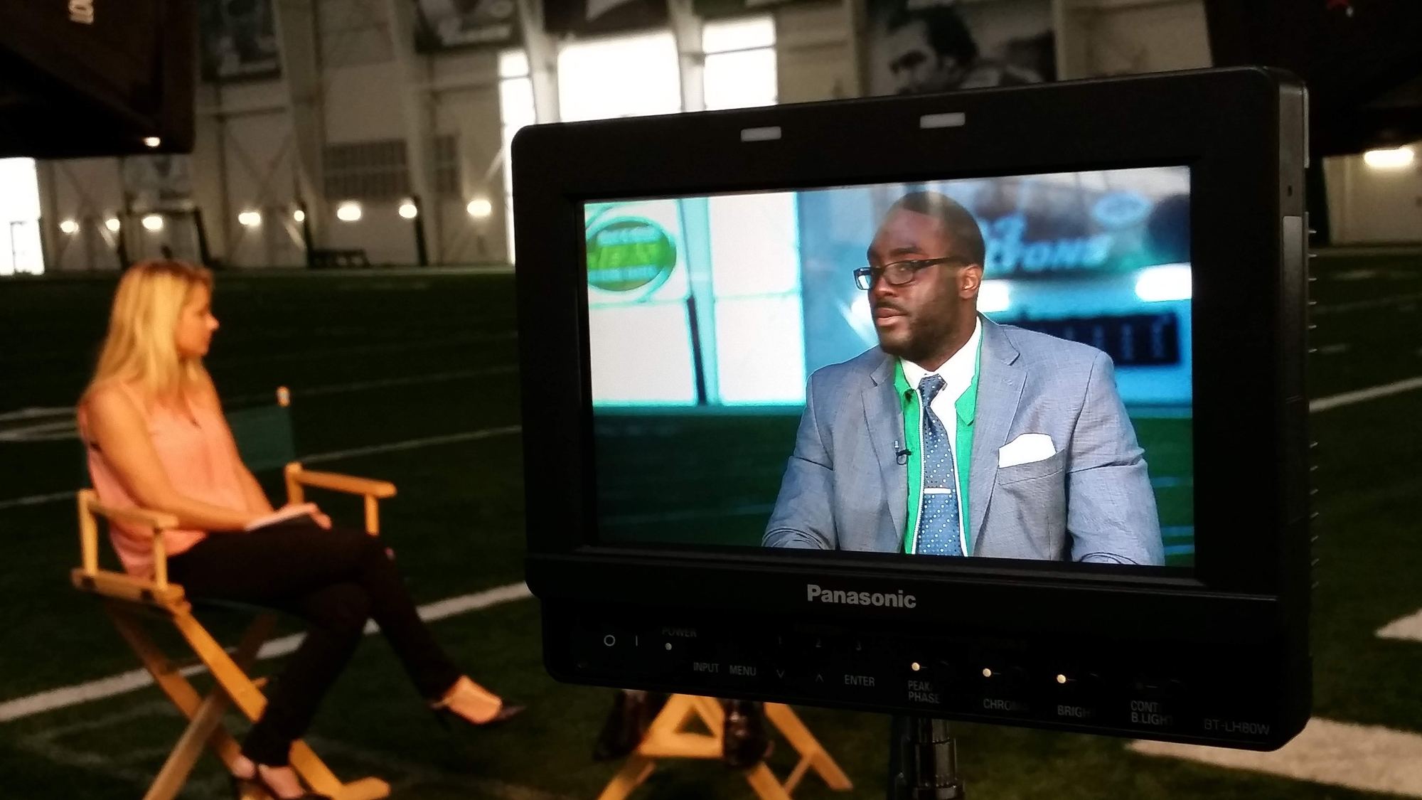 A Panasonic field monitor on the Jets indoor practice field, framing an interview subject in a grey suit.