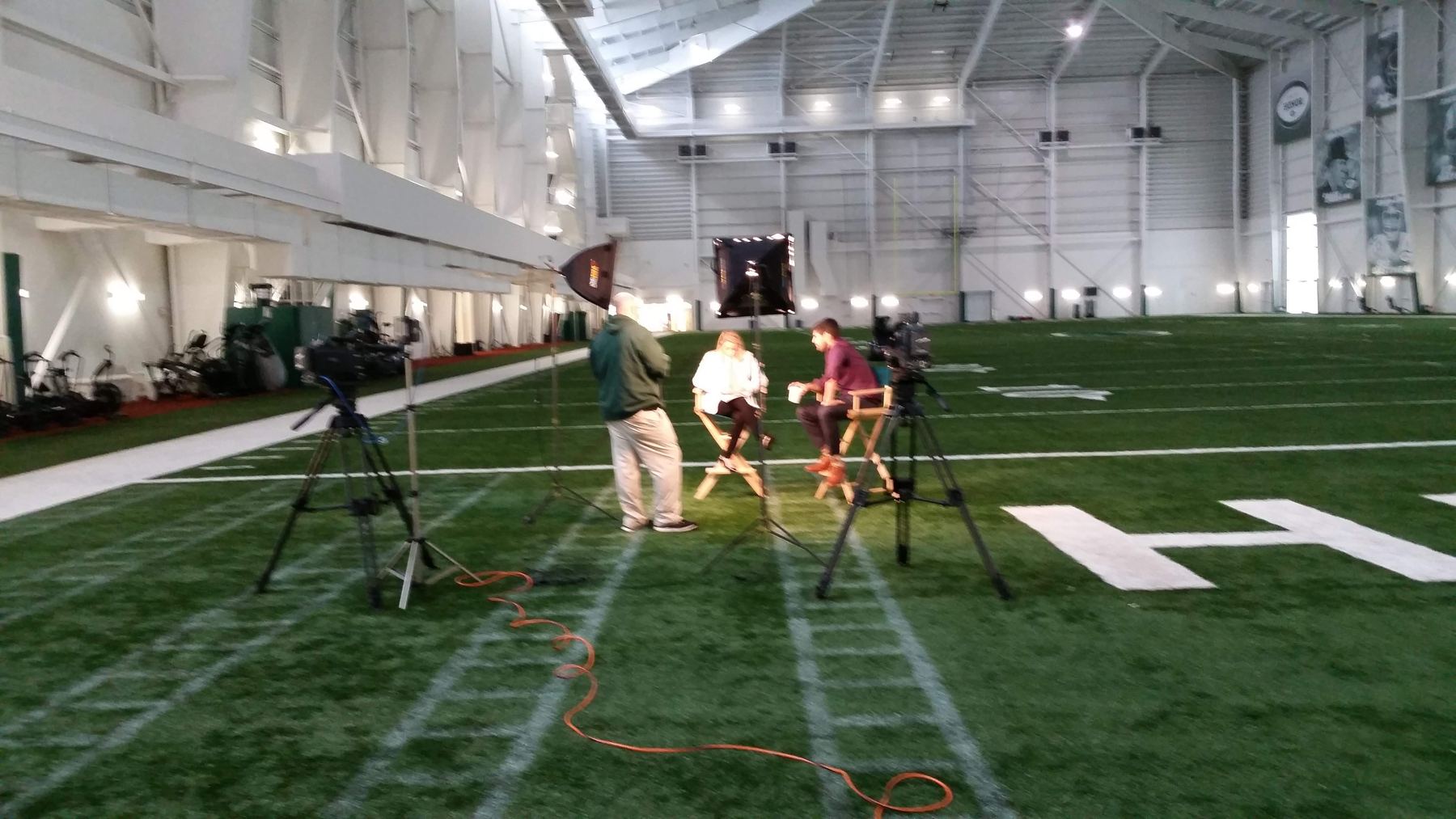 An interview set built on the Jets indoor practice field: two cameras on tripods, a light, and two people in director's chairs on the turf.