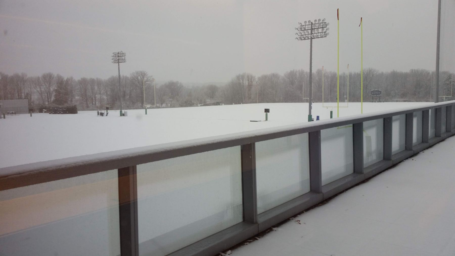 Snow covering the empty Jets outdoor practice field, seen through a window, with the goalposts and light towers in fog.