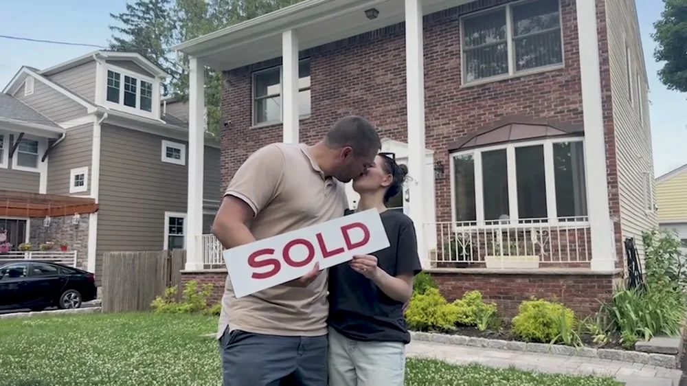 Craig and Nicole outside the home they bought, holding a SOLD sign, 2026